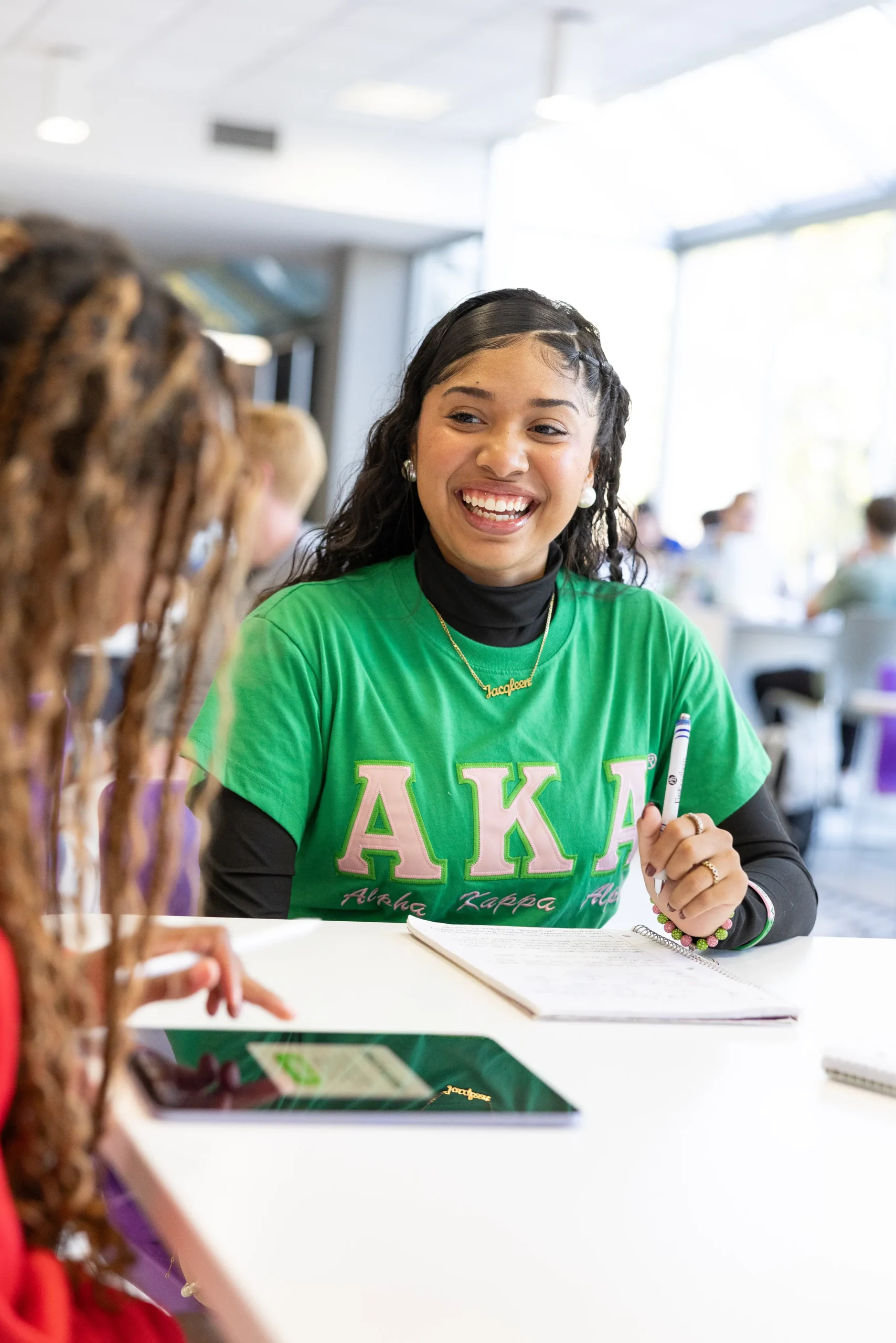 A young student wearing an AKA sorority shirt smiling and doing homework at a lunch table with friends